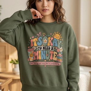 May include: An olive green crewneck sweatshirt featuring a colourful graphic print with the text "IT'S OKAY TO NEED A MINUTE" and "Your feelings are valid. Rest is productive too." The design includes a smiley face cup, sun, and books.
