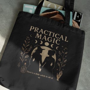 Puede incluir: Un bolso de tela negro con un estampado gráfico en beige que incluye el texto "PRACTICAL MAGIC" y "There's a little witch in all of us." El diseño muestra siluetas de dos figuras, fases lunares, elementos botánicos, una botella y una mano.