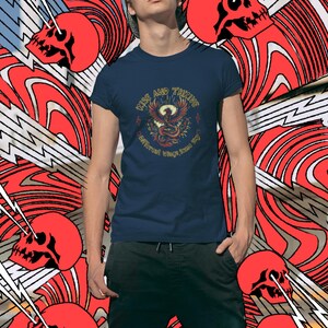 May include: Navy blue t-shirt with a graphic of a phoenix and the words "Rise and Thrive". The shirt is worn by a person in front of a red and white abstract background with red skulls and lightning bolts.