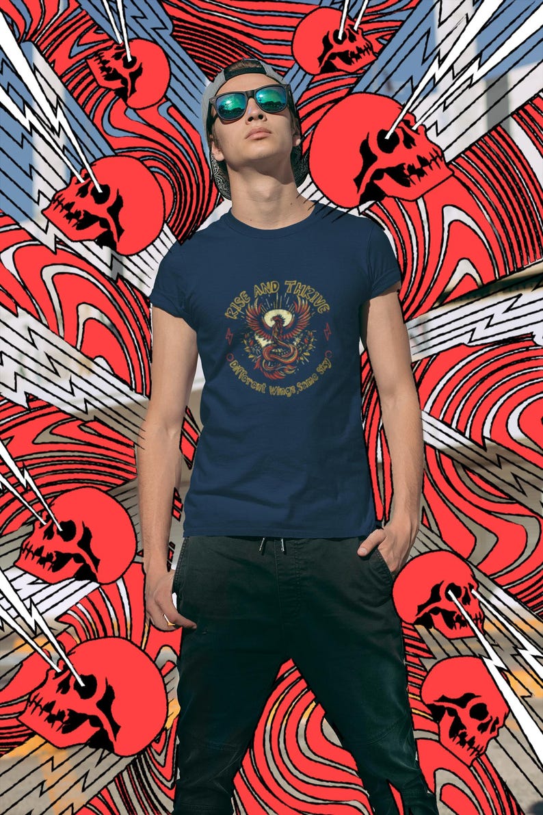 May include: Navy blue t-shirt with a graphic of a phoenix and the words "Rise and Thrive". The shirt is worn by a person in front of a red and white abstract background with red skulls and lightning bolts.