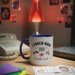 May include: White ceramic mug with a blue handle and rim, featuring the text "I TEACH KIDS to talk back" and various small illustrations. The mug sits on a wooden desk with other items, including a lava lamp and cards.