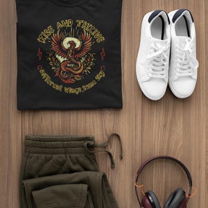 May include: A black t-shirt with a phoenix graphic and the words "Rise and Thrive." The image also includes olive green sweatpants, white sneakers, and maroon headphones, all arranged on a wooden surface. A casual outfit.