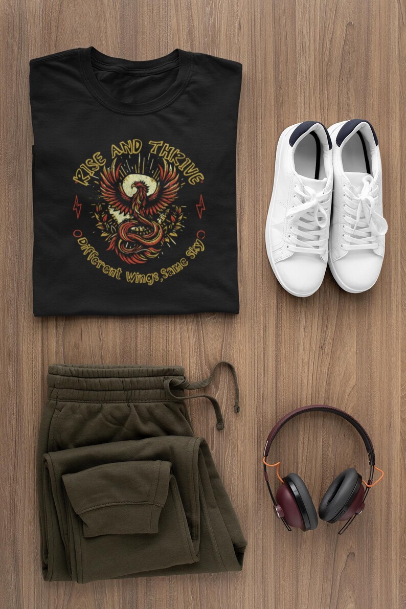 May include: A black t-shirt with a phoenix graphic and the words "Rise and Thrive." The image also includes olive green sweatpants, white sneakers, and maroon headphones, all arranged on a wooden surface. A casual outfit.