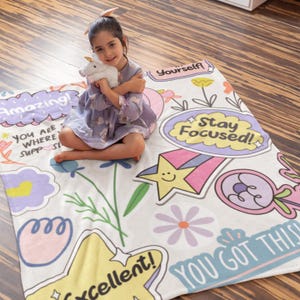 Puede incluir: Una manta blanca con parches coloridos que presentan afirmaciones positivas como "¡Increíble!" y "¡Lo tienes!" Una niña está sentada en la manta sosteniendo un unicornio de peluche.