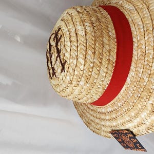 May include: A straw hat with a red band and a brown embroidered design. The hat has the text "Straw Hat" and "Monkey D. Luffy" on the side.