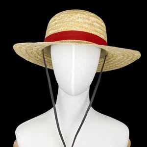 Può includere: Un cappello di paglia con un nastro rosso attorno alla corona e un cinturino in pelle marrone. Il cappello è su un manichino bianco.