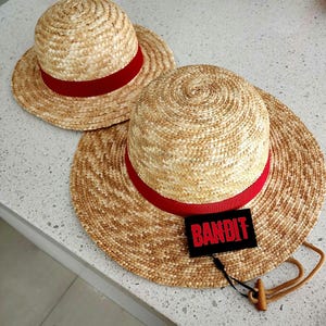 Könnte beinhalten: Zwei Strohhüte mit breiten Krempen und roten Bändern. Einer der Hüte hat einen schwarzen Aufnäher mit dem Wort "BANDIT" in roten Buchstaben. Die Hüte sind aus geflochtenem Stroh und haben eine natürliche, hellbraune Farbe.