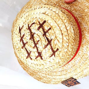 May include: A straw hat with a brown band and a brown X pattern stitched into the crown. The hat has a label on the side that reads "The Monkey D. Luffy"