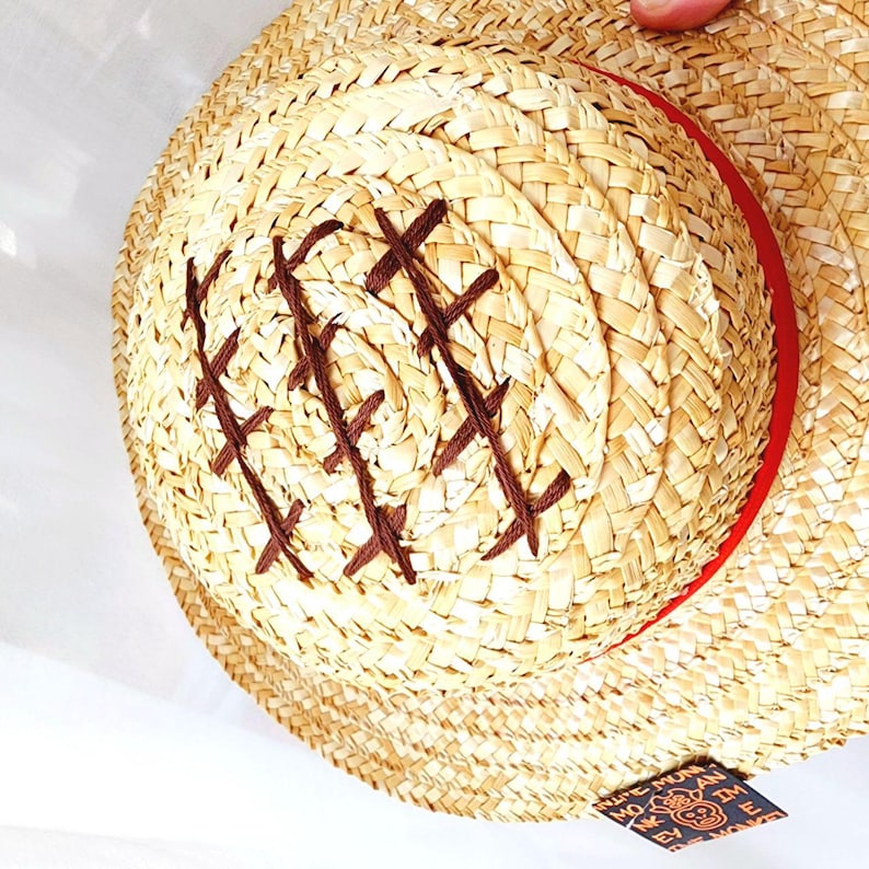 May include: A straw hat with a brown band and a brown X pattern stitched into the crown. The hat has a label on the side that reads "The Monkey D. Luffy"