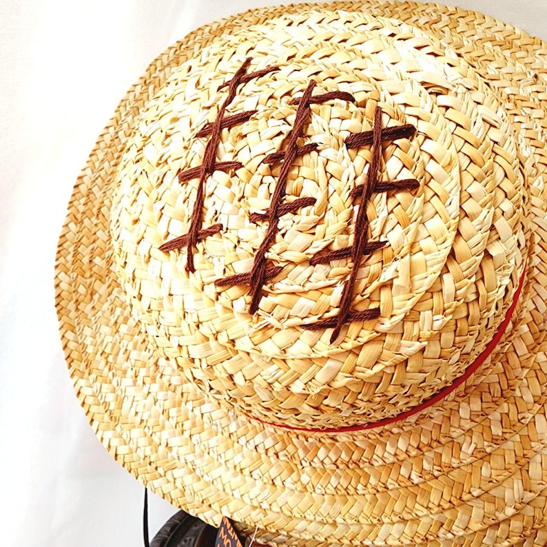 May include: A straw hat with a brown woven design and a red band around the crown. The hat has a wide brim and is made of natural materials.