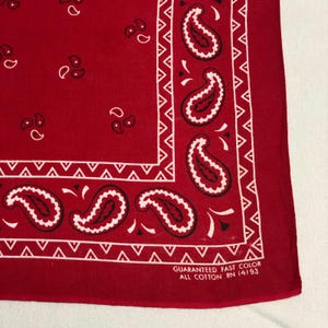 May include: A red bandana featuring white paisley patterns and a white border. The text "GUARANTEED FAST COLOR ALL COTTON RN 14193" is printed on the bandana. The bandana is made of all cotton.