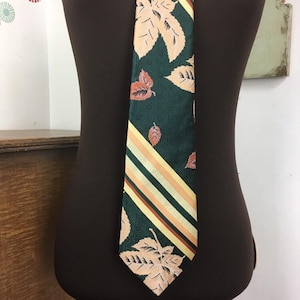 May include: A dark green tie featuring a leaf pattern and diagonal stripes in yellow, orange, and beige. The tie is displayed on a mannequin, highlighting its design and colours.