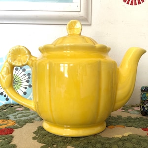 May include: A bright yellow ceramic teapot with a lid and spout. The handle is adorned with a lemon design. The teapot has a ribbed body and sits on a round base. A charming kitchenware item.