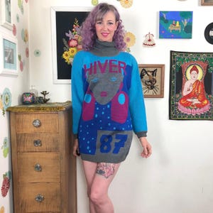 Könnte beinhalten: Ein langärmeliges Strickkleid in Grau und Blau mit hohem Rollkragen. Die Vorderseite zeigt eine stilisierte Grafik eines Gesichts in Grau, darüber das Wort HIVER in Rot und am unteren Saum die Zahl 87 in Blau.
