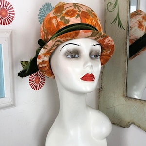 May include: A vintage cloche hat with a floral pattern in orange and peach tones. The hat features a dark green velvet band and a bow on the side. The hat is displayed on a mannequin head.