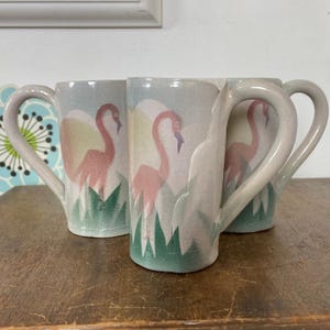 May include: Three ceramic mugs with flamingo designs. Each mug features a pink flamingo with a yellow and white background, green grass, and a light blue sky. The mugs have large, curved handles and are set on a wooden surface.