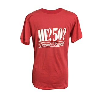 May include: Red t-shirt with white text that reads "ME? 50? I Demand A Recount!" The shirt has short sleeves and a crew neck. The text is in a bold, sans-serif font.