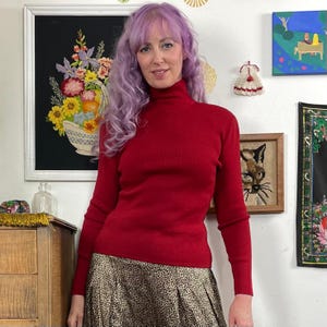 Puede incluir: Una mujer con un jersey rojo de cuello alto y una falda con estampado de leopardo. El jersey tiene mangas largas y un corte ajustado. La falda es plisada y llega a las rodillas. La mujer tiene el pelo morado y sonríe.