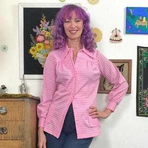 May include: A pink and white gingham button-up shirt with long sleeves and a collared neckline. The shirt has two front pockets and is paired with blue jeans. The shirt is a vintage style.