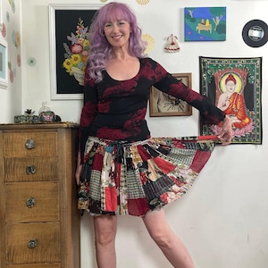 Puede incluir: Una mujer con una blusa negra de manga larga con estampados florales rojos y una falda patchwork. La falda presenta una variedad de telas en rojo, verde y blanco. El fondo incluye obras de arte y una cómoda de madera.