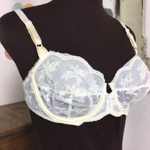 May include: Ivory-colored bra with floral lace overlay. The bra features underwire support, adjustable straps, and a delicate scalloped trim along the edges. The bra is displayed on a mannequin.