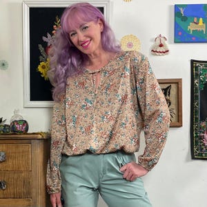 May include: A tan floral print blouse with long sleeves and a keyhole neckline. The blouse is paired with light green pants. The woman in the image has purple hair and is smiling. The background includes framed artwork and decorative items.