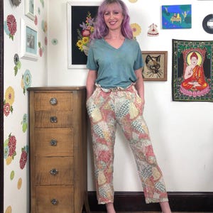Könnte beinhalten: Eine Person trägt ein türkisfarbenes V-Ausschnitt-Shirt und eine gemusterte Hose. Die Hose hat ein Patchwork-Design in Rosa, Gelb und Blau. Links ist eine Holzkommode. Der Hintergrund umfasst gerahmte Kunstwerke und Wandaufkleber.