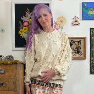 May include: Cream-colored, hand-knitted sweater with a floral pattern. The sweater features raised, textured floral details across the front and sleeves. The neckline and cuffs have a ribbed design. The model is wearing a floral patterned skirt.