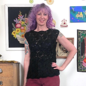 May include: A black, sleeveless top with a textured, openwork design. The top is worn with burgundy pants. The background includes framed artwork and decorative wall hangings. The woman has purple hair and a tattoo on her arm.