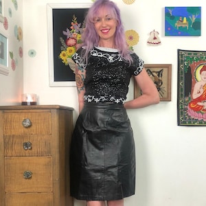 May include: A black short-sleeved top featuring dense black sequins and white beaded floral embroidery around the neckline and sleeves, paired with a high-waisted black leather skirt that falls to the knee with a smooth, semi-gloss finish.