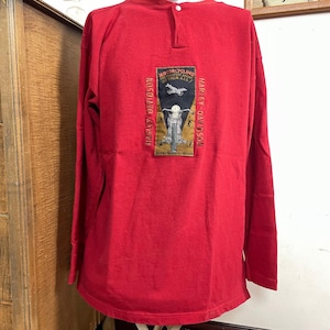 May include: Red long-sleeve shirt with a small button closure at the back of the neck. The shirt features a Harley-Davidson patch on the back with a motorcycle graphic and the text "Motorcycling The Greatest Sport Of Them All."