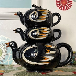 May include: A stacked black ceramic teapot with a duck design. The teapot has three sections, each with a duck head spout and a handle. The design includes white and orange accents. A small duck figurine sits on the lid.