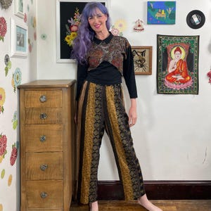 Puede incluir: Una camisa negra de manga larga con una sobrecapa corta con cuello y estampado en tonos marrón, naranja y negro. El conjunto incluye pantalones holgados de cintura alta con paneles verticales de patrones florales en color mostaza y negro.