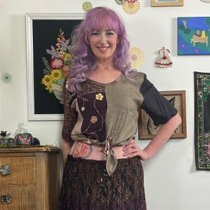 May include: A short-sleeved top featuring a patchwork design with a dark brown floral panel, a solid olive green panel, and a black sleeve. The front of the top includes a tie-front detail at the hem. The garment is paired with a long, dark brown skirt.