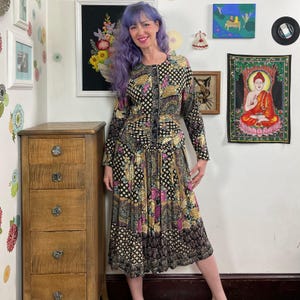 May include: A long-sleeved dress featuring a black base with a multi-coloured pattern of gold, pink, and white floral and geometric shapes. The dress has a fitted bodice, a button-down front, and a pleated skirt that falls below the knee.