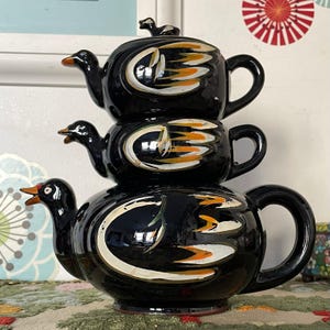 May include: A three-tiered black ceramic teapot with a duck theme. Each section features a duck head spout and a handle. The design incorporates white and orange details. A small duck figurine is on the lid.