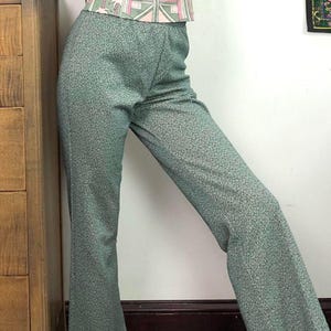 May include: Green, textured flared trousers. The trousers have a subtle pattern and a high waist. The top of the trousers are visible, with a patterned top in pink, green, and white. The trousers are made of a soft, comfortable material.