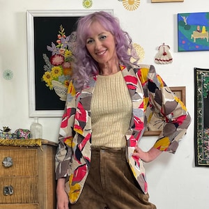May include: A colorful blazer with a unique pattern of red, yellow, and gray shapes. The blazer is worn over a ribbed, tan-colored top and brown pants. The background includes framed artwork and decorative elements.