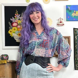 May include: A woman wearing a patterned blouse with a black leather belt and a light blue skirt. The blouse features a colorful feather-like design in shades of blue, pink, and green. The belt cinches the waist, and the skirt has a textured appearance.