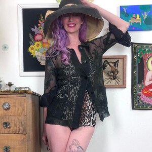 May include: A black sheer lace jacket with a button-up front and three-quarter sleeves. The jacket is paired with leopard print shorts and a wide-brimmed sun hat. The hat is black with a tan underside. The outfit is accessorized with a tattoo on the leg.