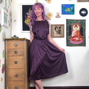 May include: A plum-colored halter dress with a gathered neckline and a flowing skirt. The dress has a cinched waist and is knee-length. The model is holding the skirt out, showcasing the dress's shape. The background includes framed art and a wooden dresser.