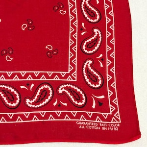 May include: A bright red bandana featuring a white paisley pattern and a geometric border. The fabric includes printed text in the corner that reads: GUARANTEED FAST COLOR ALL COTTON RN 14193. The design consists of repeating teardrop shapes.