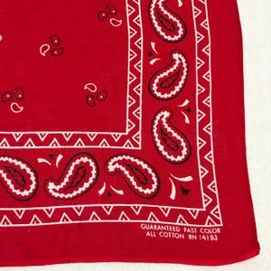May include: Red bandana with a white paisley pattern and border. The bandana features a repeating paisley design along the edges and smaller paisley shapes scattered across the field. The text "GUARANTEED FAST COLOR ALL COTTON RN 14193" is printed on the bottom right corner.