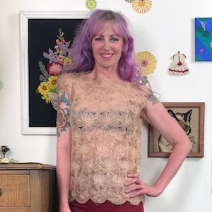 May include: A beige, short-sleeved top with a floral pattern. The top has a scalloped hemline and is worn with a dark undergarment. The person in the image is smiling and has purple hair.