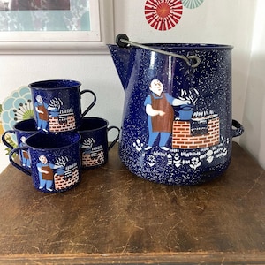 May include: A dark blue speckled enamel coffee pot and three matching mugs. Each piece features a painted illustration of a person in a brown apron cooking at a brick stove. The coffee pot has a metal handle and a spout, while the mugs have side handles.