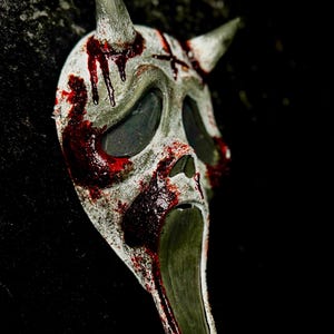 May include: A white, elongated mask featuring two pointed horns at the top, large hollow eye sockets with dark mesh, and an open, stretched mouth. The surface is distressed with splatters of deep red paint resembling blood across the face.