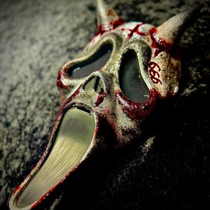 May include: A textured, off-white mask with two pointed horns, featuring dark eye sockets and a long, tapered chin. The surface is splattered with red paint resembling blood, and the number 666 is written in red on the right side of the forehead.