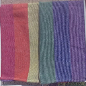 Two folded handwoven rainbow pride hand towels hang side by side, one showing red, orange, and yellow stripes, the other green, blue, and purple stripes. Each stripe is just shy of 3 inches wide.