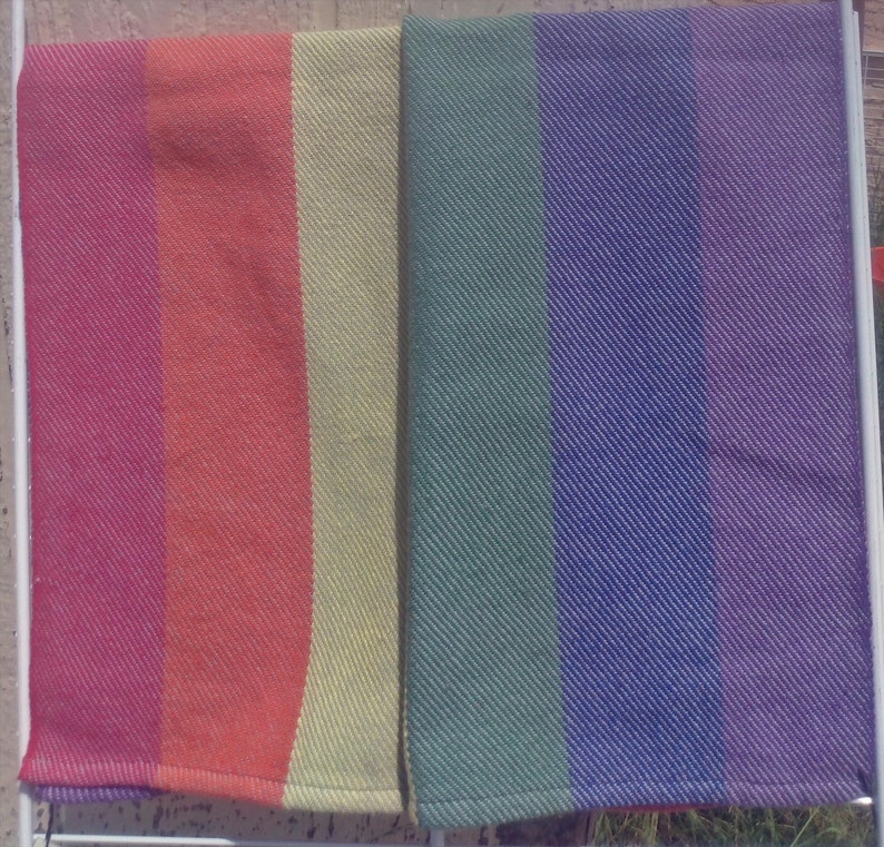 Two folded handwoven rainbow pride hand towels hang side by side, one showing red, orange, and yellow stripes, the other green, blue, and purple stripes. Each stripe is just shy of 3 inches wide.