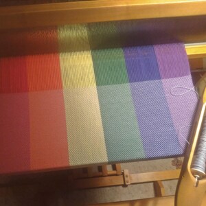 The fabric is shown being woven on a four-harness Harrisville loom. The shuttle holding the light silver weft yarn sits at an angle on the lower right corner of the fabric.